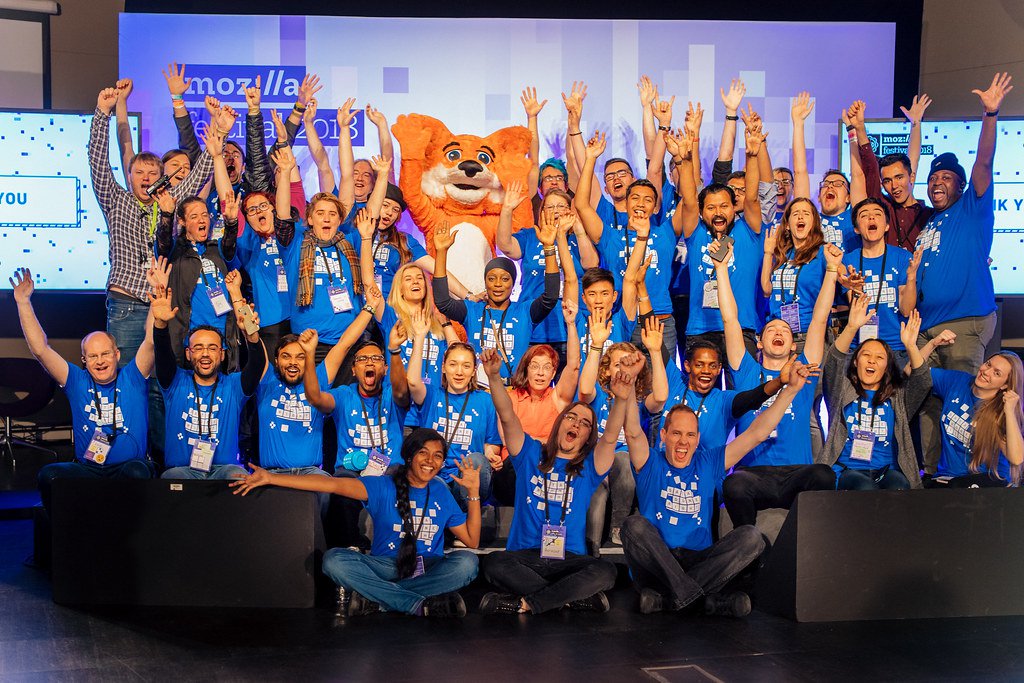 A costumed cartoon fox raises its hands in celebration while surrounded by a group of MozFest Volunteers wearing MozHelp t-shirts.
