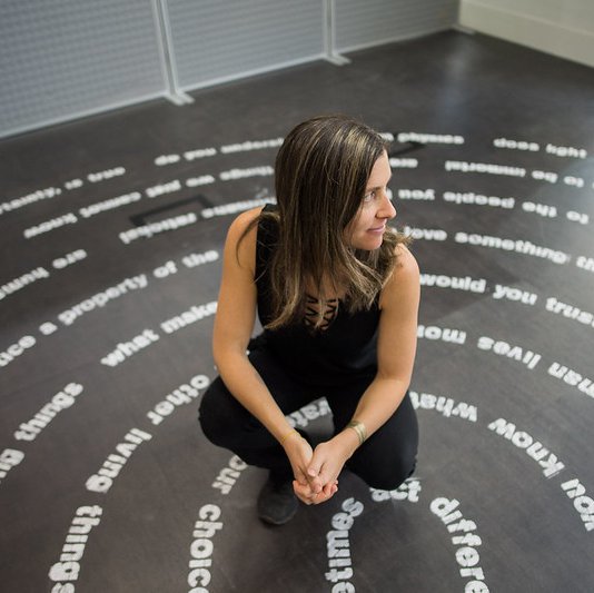 An artist, sitting with one of their art pieces, of various moral questions in the shape of a labyrinth on the floor.