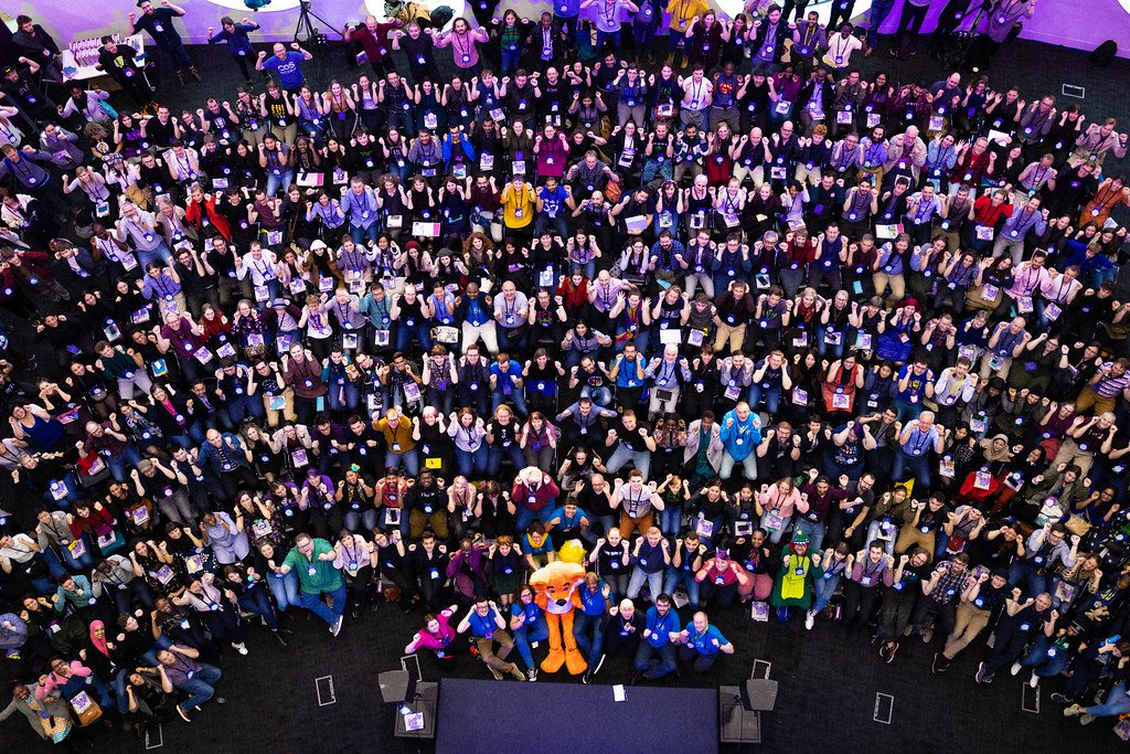 An overhead shot of the audience at the MozFest opening circle in 2019 showing hundreds of people looking up at the camera while seated in a semicircle in front of a small stage.