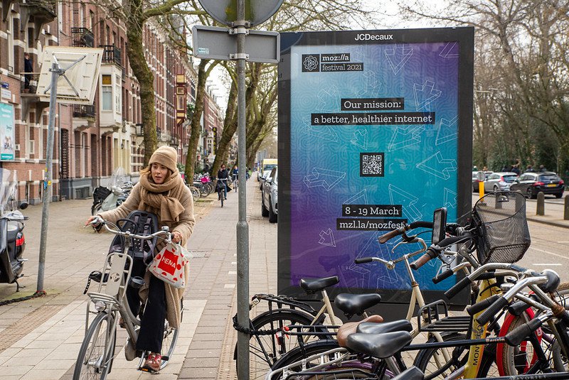 On a street in Amsterdam, digital signage announces MozFest 2021, while people on bikes zip by