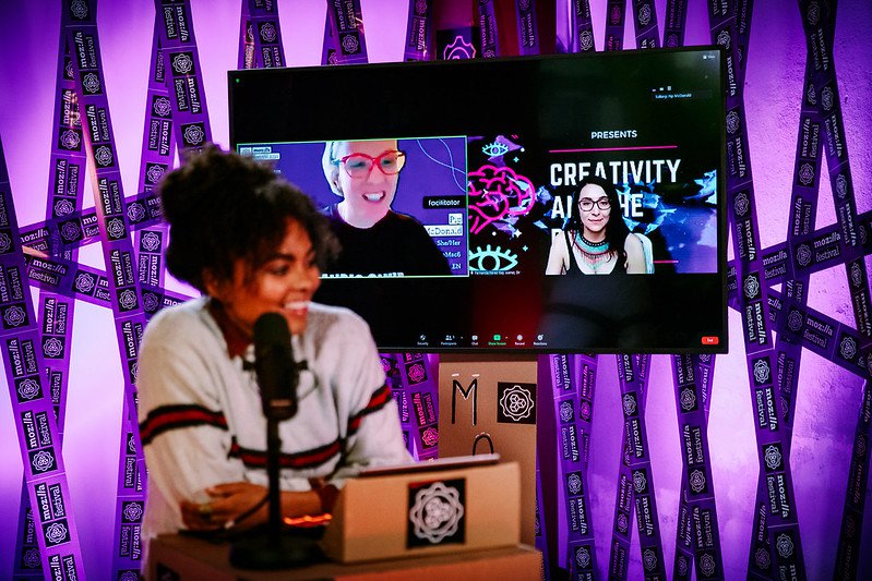 A presenter stands at a lectern, in front of a television screen that has a video conference with two people.