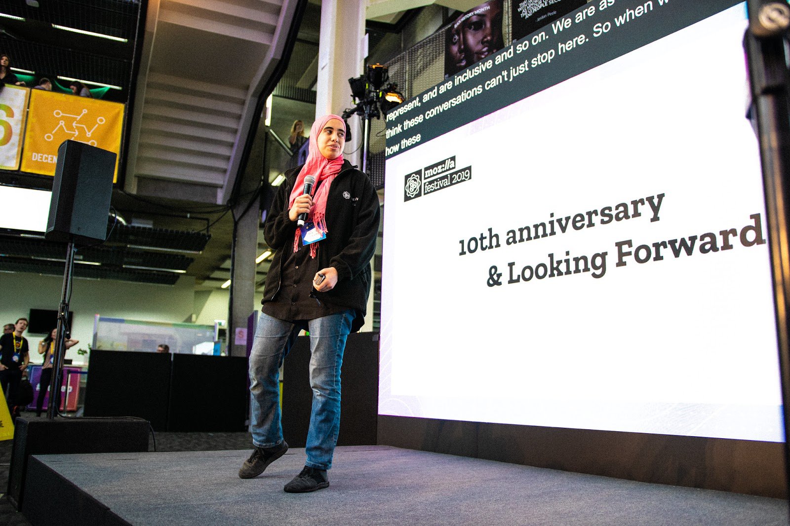 Stage at an indoors event with a person holding a microphone looking back at the screen