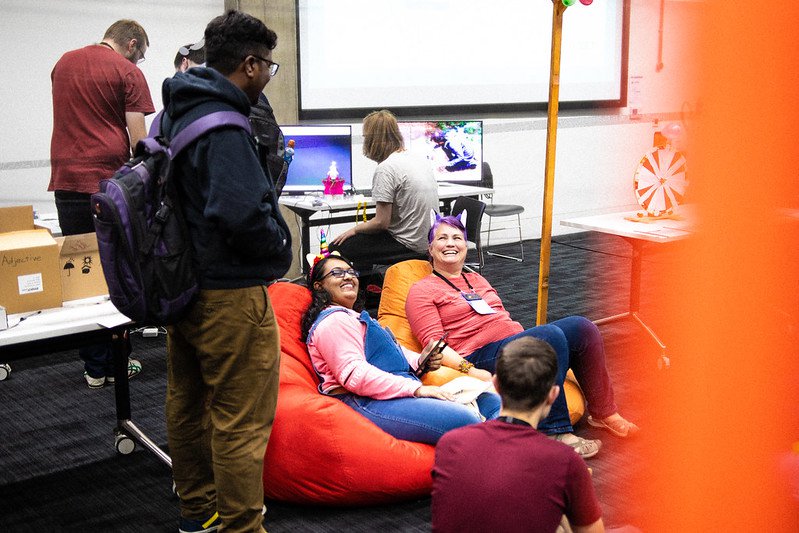 Two people sitting on a large, orange bean bag and smiling up at a person standing next to them with a backpack on, faced away from the camera.