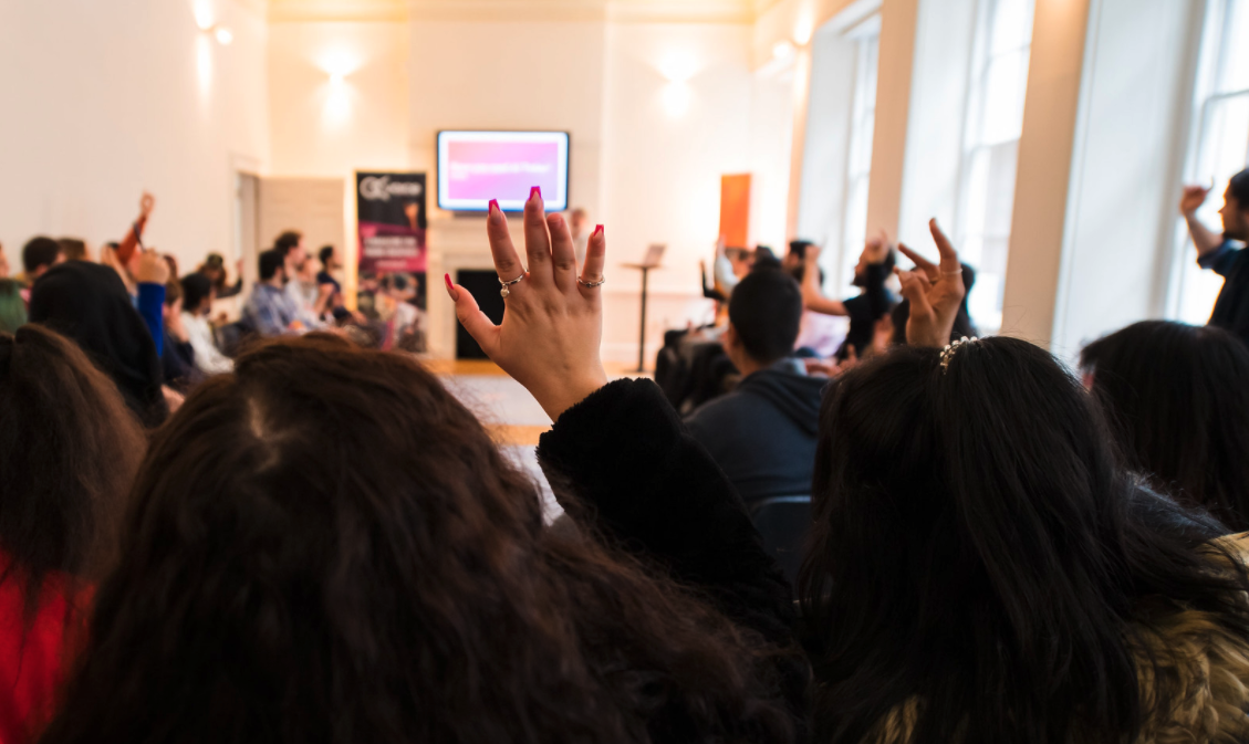 A photo captured from the back of a crowded room with people's hands raised towards the speaker at the front of the room