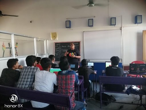 Photo taken from the back of a classroom with white walls in which all the people are looking at the speaker standing at the front of the room.