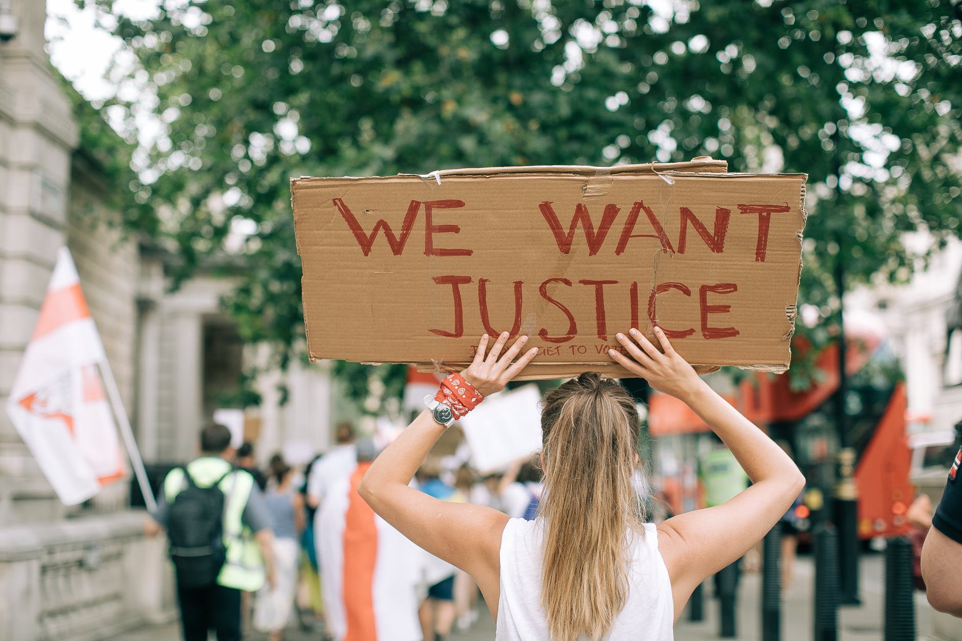 A person with long hair pulled into a ponytail facing away from the camera, holding a sign above their head that reads "We Want Justice"