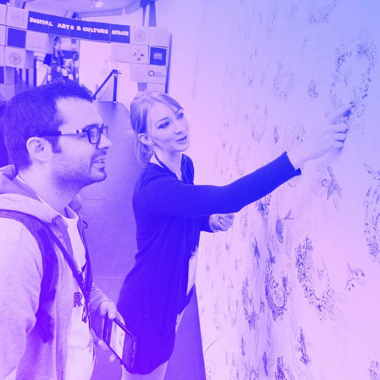 Two people stand next to a piece of art on the wall, one of the people is pointing to the point and explaining the work.
