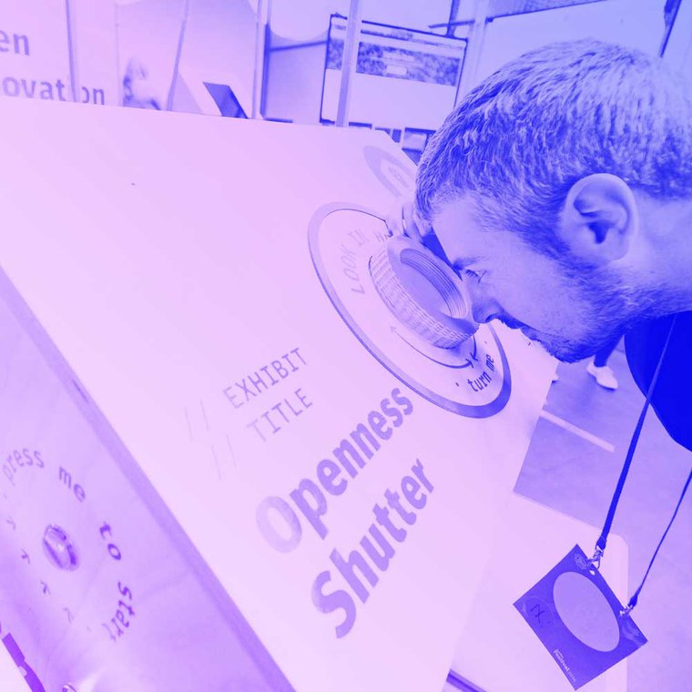A person stands at an installation looking into a lens. The title of the installation is printing onto the side and reads 'Openness Shutter'