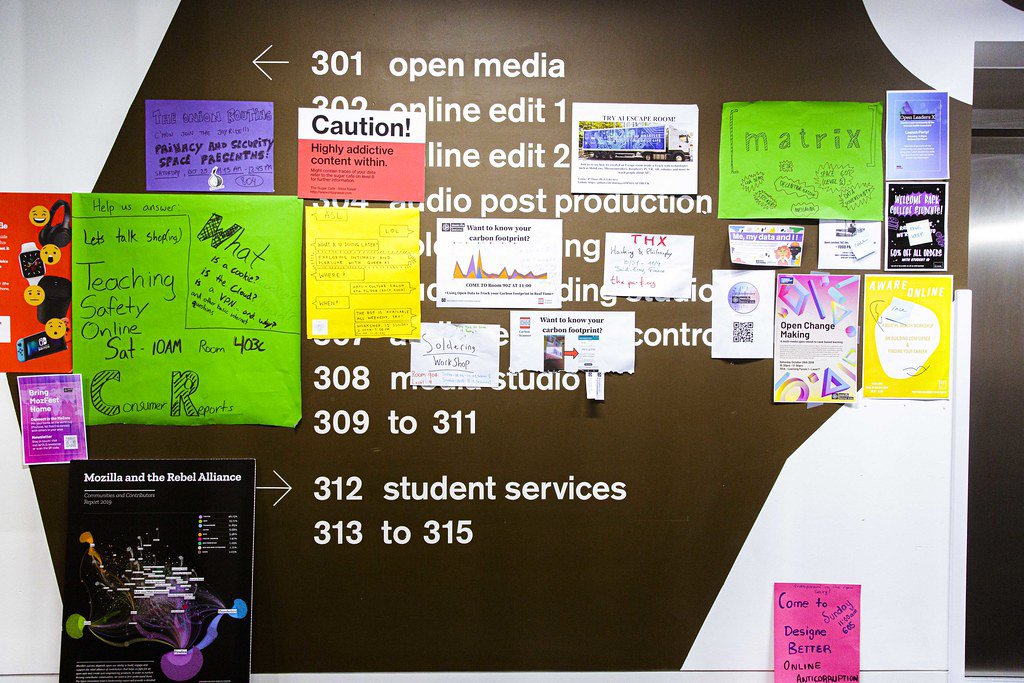 An image of a wall that’s been painted brown with white lettering to list a directory of rooms on the 3rd floor of Ravensbourne University in London with brightly multi-colored MozFest session posters stuck on top of it sharing session titles like “Teaching safety online,” “Caution! Highly addictive content within,” and “Open change making.”