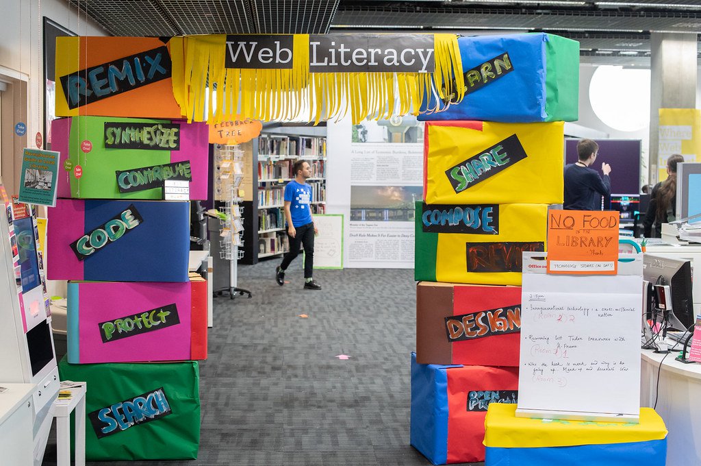 An image of cardboard boxes stacked and stuck together to make a squared arch with each box covered in brightly colored construction paper signs with words like, “Web Literacy,” “Learn,” and “Remix.”