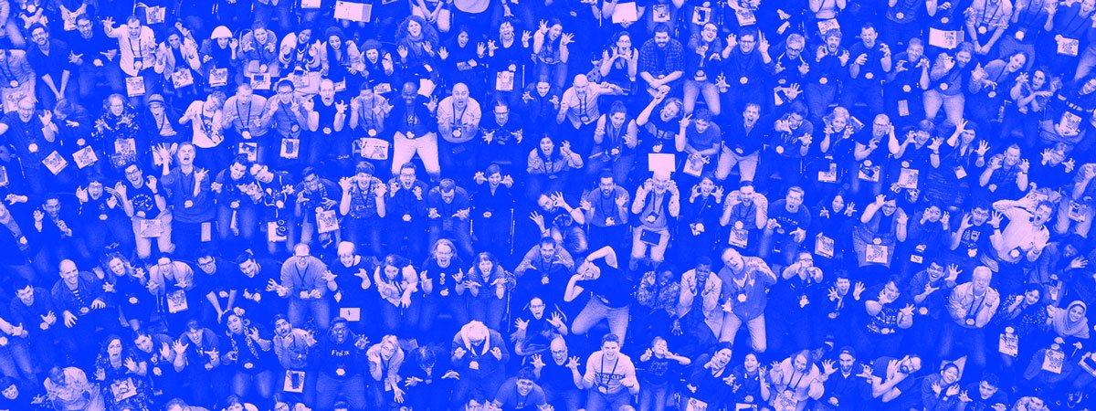 A group photo, taken from above, of hundreds of MozFest attendees sitting in chairs arranged in a semi-circle in front of a small plenary stage.